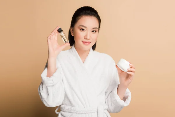 Sourire belle asiatique femme en peignoir tenant crème cosmétique et huile sur fond beige — Photo de stock