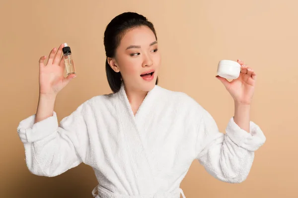 Beautiful asian woman in bathrobe holding cosmetic cream and oil with open mouth on beige background — Stock Photo