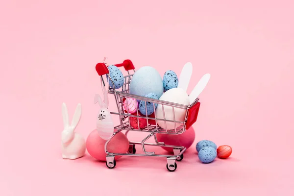 Ovos de páscoa coloridos e coelhos decorativos no carrinho no fundo rosa — Fotografia de Stock