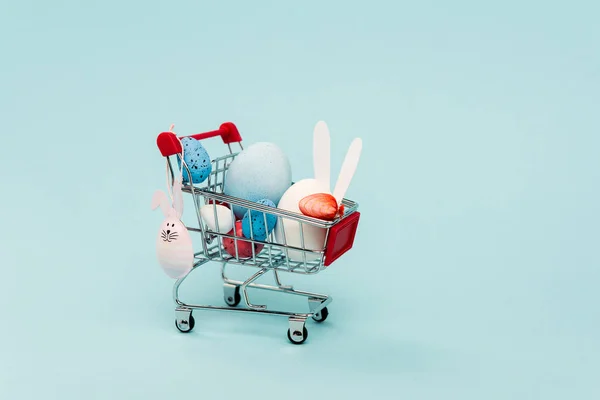 Colorful easter eggs and decorative rabbits in cart on blue background — Stock Photo