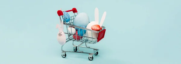 Bunte Ostereier und Zierhasen im Einkaufswagen auf blauem, panoramischem Foto — Stockfoto