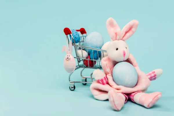 Carrinho com ovos de páscoa coloridos e coelhos decorativos em fundo azul — Fotografia de Stock