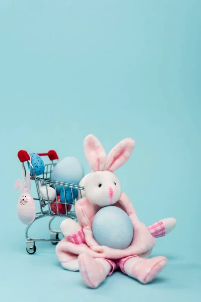 Carro con huevos de Pascua coloridos y conejitos decorativos sobre fondo azul - foto de stock