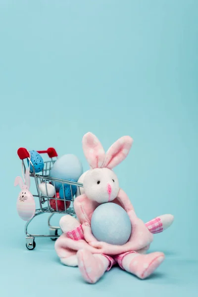 Carro con huevos de Pascua coloridos y conejitos decorativos sobre fondo azul - foto de stock