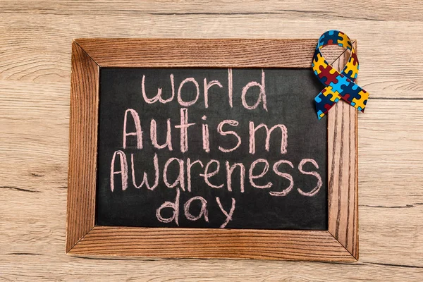 Top view of board with world autism awareness day lettering and ribbon on wooden background — Stock Photo