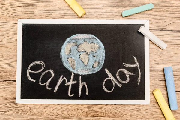 Top view of black board with earth day lettering and pieces of chalk on wooden background — Stock Photo