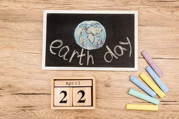 Top view of board with earth day lettering, pieces of chalk and calendar with 22 april inscription on wooden background — Stock Photo