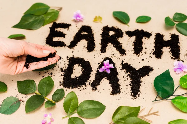Cropped view of handful of ground in female hand, earth day lettering with leaves and buds on beige — Stock Photo