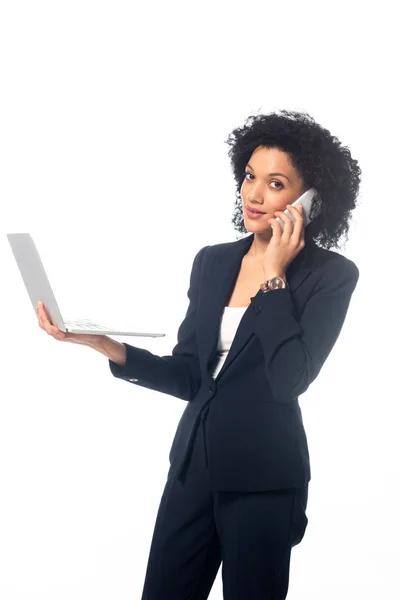 Exitosa mujer de negocios afroamericana mirando a la cámara, sosteniendo la computadora portátil y hablando por teléfono aislado en blanco - foto de stock