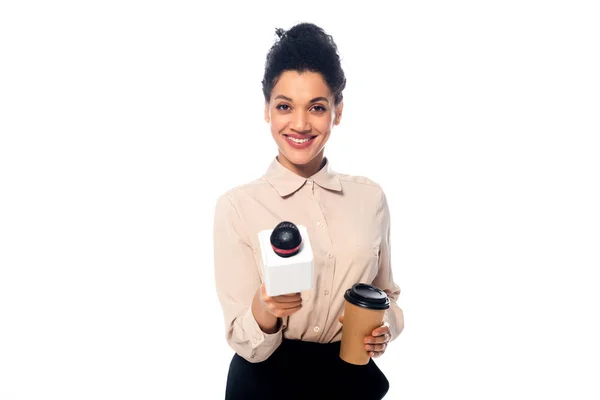 Vista frontal do jornalista afro-americano com copo descartável de café sorrindo e mostrando microfone isolado em branco — Fotografia de Stock