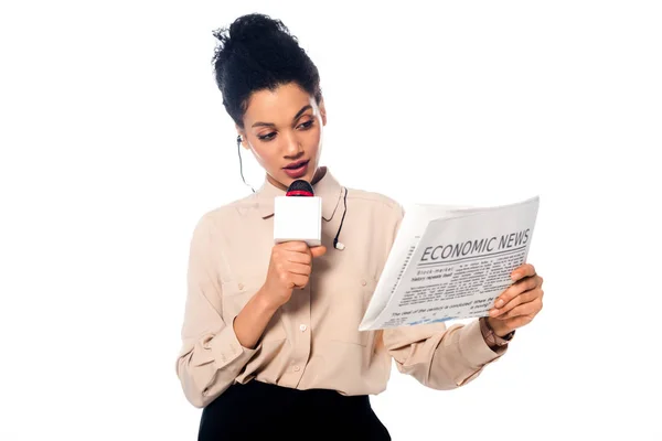 Periodista afroamericano hablando en micrófono y mirando el periódico con letras de noticias económicas aisladas en blanco - foto de stock