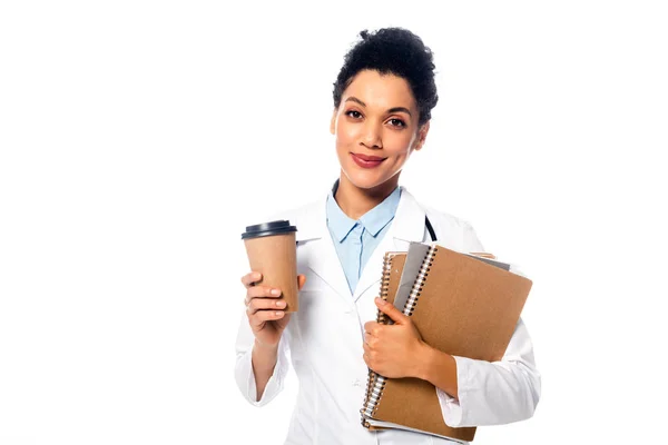 Médecin afro-américain avec tasse jetable de café, cahiers et dossier regardant la caméra isolée sur blanc — Photo de stock