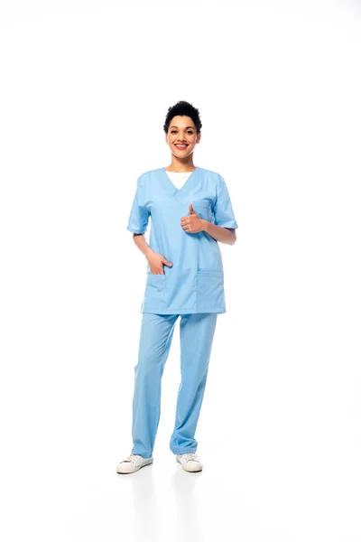 Vista a figura intera di infermiera afro-americana con mano in tasca e pollice in su sorridente e guardando la fotocamera su bianco — Foto stock
