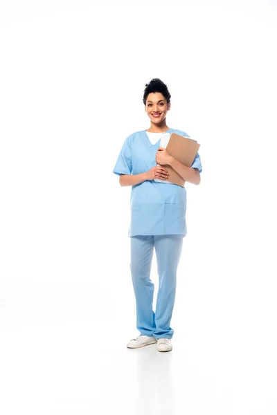 Full length view of african american nurse with papers smiling and looking at camera on white — Stock Photo