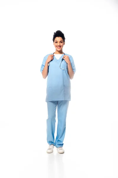 Vista completa de enfermera afroamericana con estetoscopio sonriendo sobre fondo blanco - foto de stock