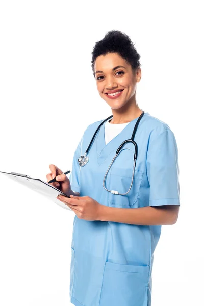 Enfermera afroamericana con estetoscopio escribiendo en portapapeles y sonriendo aislada en blanco - foto de stock