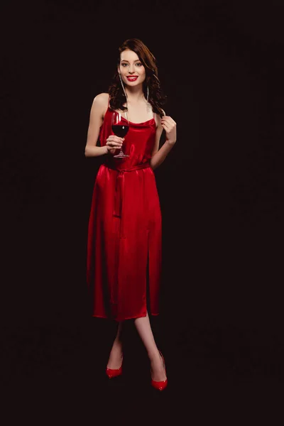 Longitud completa de hermosa mujer en vestido sosteniendo copa de vino tinto y sonriendo a la cámara aislada en negro - foto de stock