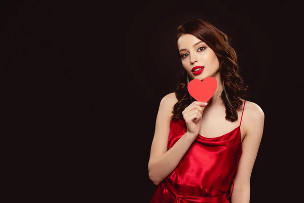 Jolie jeune femme en robe rouge tenant du papier en forme de coeur et regardant la caméra isolée sur noir — Photo de stock