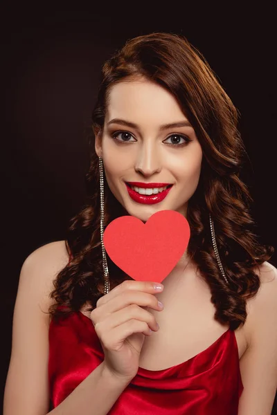 Attractive brunette woman with red lips smiling at camera while holding paper heart isolated on black — Stock Photo