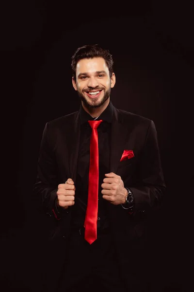 Hombre guapo en traje negro y corbata roja sonriendo a la cámara aislada en negro - foto de stock
