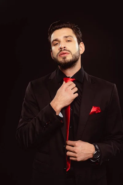 Elegante hombre ajustando corbata roja y mirando a la cámara aislada en negro - foto de stock
