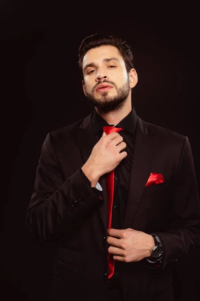 Elegant man adjusting red tie and looking at camera isolated on black — Stock Photo