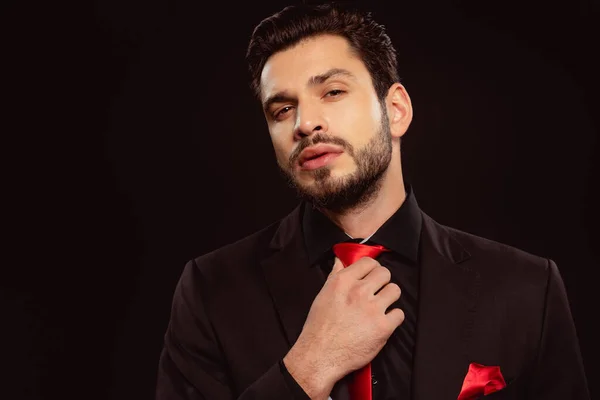 Elegante hombre mirando a la cámara y ajustando la corbata roja aislada en negro - foto de stock
