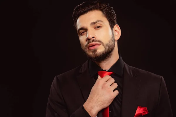 Elegant man looking at camera and adjusting red tie isolated on black — Stock Photo