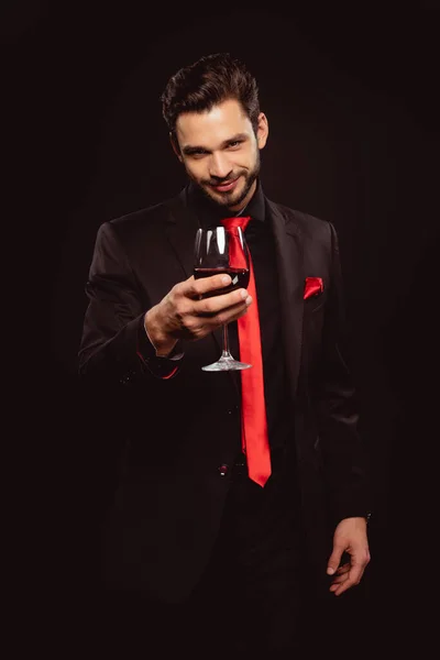 Вибірковий фокус красивого чоловіка тримає келих червоного вина і дивиться на камеру ізольовано на чорному — стокове фото