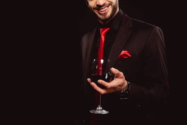 Vue recadrée de l'homme élégant souriant tout en tenant un verre de vin rouge isolé sur noir — Photo de stock