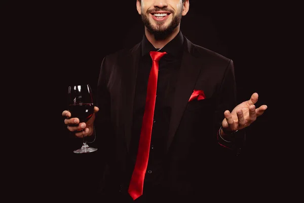 Обрезанный вид улыбающегося мужчины в костюме, жестикулирующего, держа бокал красного вина, изолированного на черном — стоковое фото