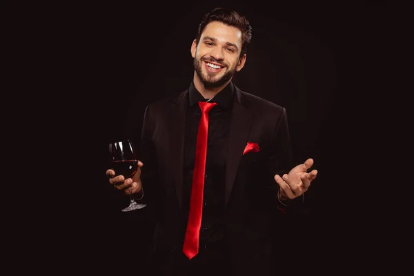 Hombre guapo en traje y corbata roja sosteniendo copa de vino y sonriendo a la cámara aislada en negro - foto de stock