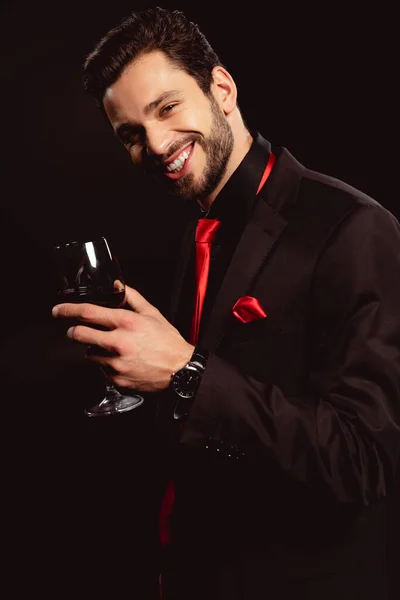 Handsome man in formal wear holding glass of red wine and smiling at camera isolated on black — Stock Photo