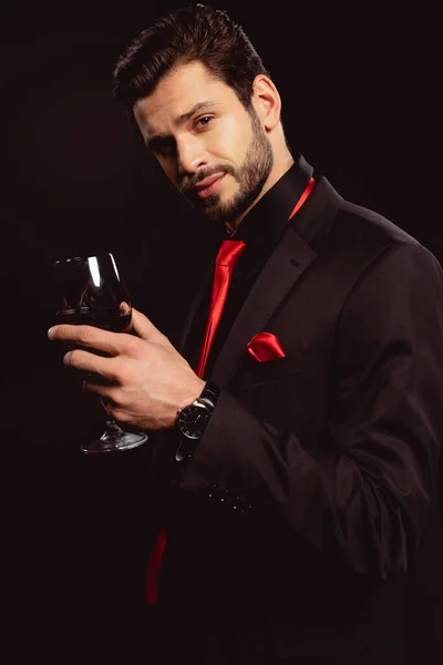 Jovem elegante segurando copo de vinho tinto e olhando para a câmera isolada em preto — Fotografia de Stock