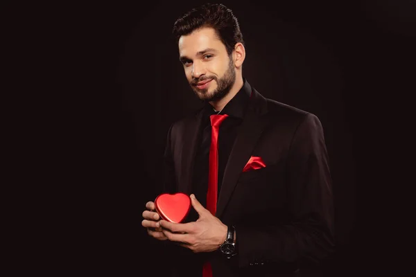Elegante hombre sosteniendo caja de regalo en forma de corazón y mirando a la cámara aislada en negro - foto de stock