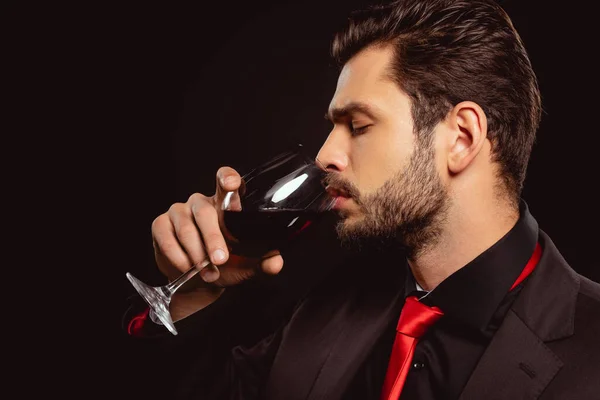 Vista lateral de homem bonito em terno beber vinho tinto isolado em preto — Fotografia de Stock