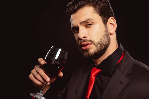 Homem bonito em desgaste formal e gravata vermelha olhando para a câmera enquanto segurando vidro de vinho tinto isolado em preto — Fotografia de Stock