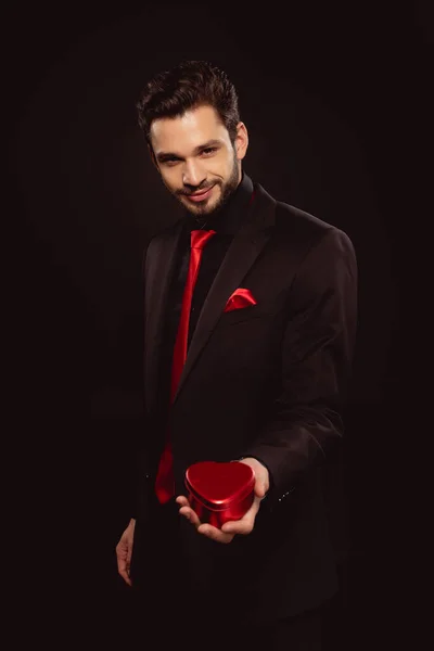 Elegante hombre de traje sosteniendo caja de regalo en forma de corazón y mirando a la cámara aislada en negro - foto de stock