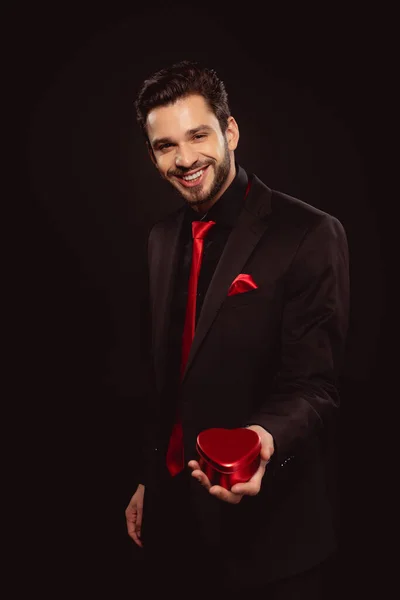 Hombre sonriente en ropa formal sosteniendo caja de regalo en forma de corazón y mirando a la cámara aislada en negro - foto de stock