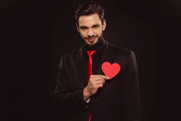 Handsome man holding paper heart and looking at camera isolated on black — Stock Photo