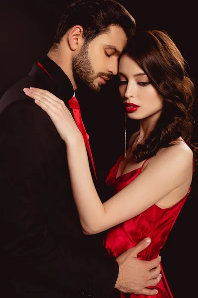 Vue latérale de belle femme en robe embrassant élégant petit ami isolé sur noir — Photo de stock