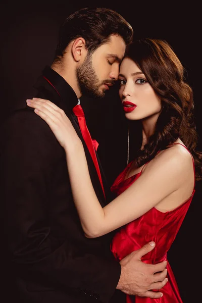 Vista lateral de la mujer elegante mirando a la cámara mientras abraza novio guapo aislado en negro - foto de stock