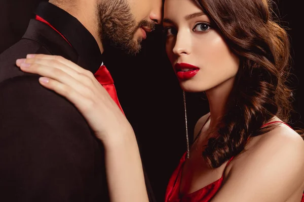 Vista lateral de hermosa chica con labios rojos mirando a la cámara mientras abraza novio aislado en negro - foto de stock