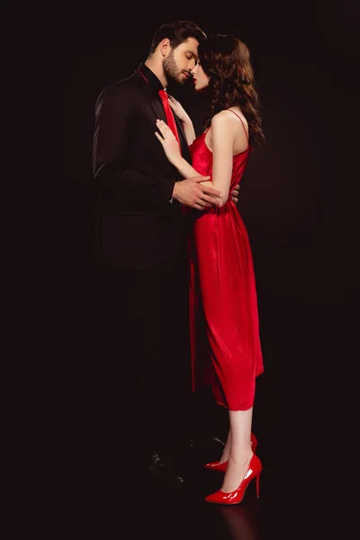 Vista lateral del hombre guapo abrazando hermosa novia en vestido rojo aislado en negro - foto de stock