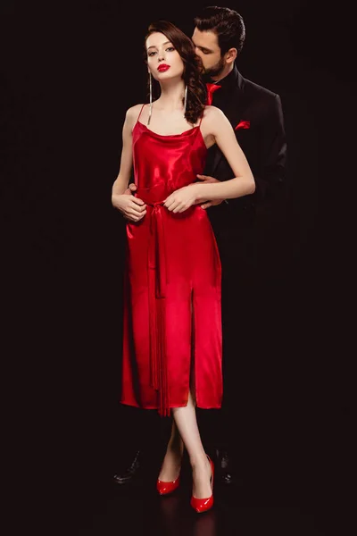 Comprimento total de mulher elegante em vestido vermelho olhando para a câmera perto namorado bonito em terno isolado em preto — Fotografia de Stock