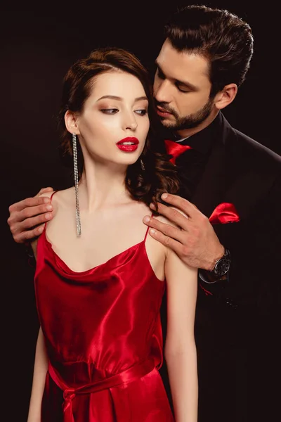 Elegante uomo in abito formale abbracciando bella fidanzata in abito rosso isolato su nero — Foto stock