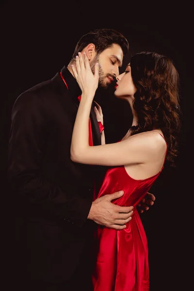 Vista lateral de mujer elegante en vestido rojo besando novio guapo en traje aislado en negro - foto de stock