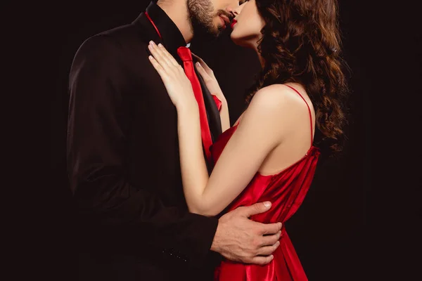 Corte vista de elegante homem no formal desgaste beijando e abraçando namorada no vermelho vestido isolado no preto — Fotografia de Stock