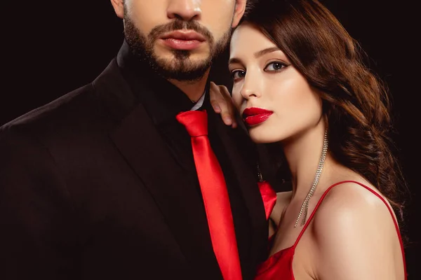 Elegante mujer con labios rojos mirando a la cámara escuchar novio en ropa formal aislado en negro - foto de stock