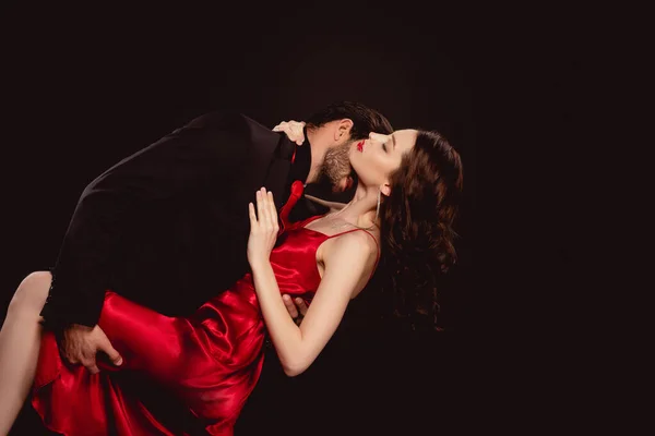 Вид сбоку элегантного мужчины, целующего в шею чувственную подругу в красном платье, изолированном от черного — стоковое фото